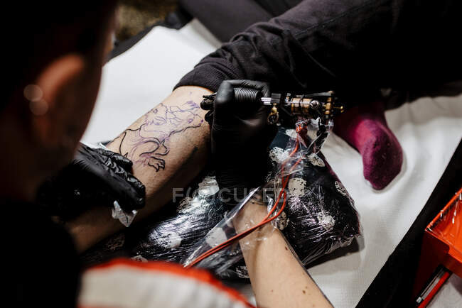 De arriba recortado hombre irreconocible con el uso de la máquina de tatuaje para hacer tatuaje en la pierna del cliente de la cosecha durante el trabajo en el salón - foto de stock