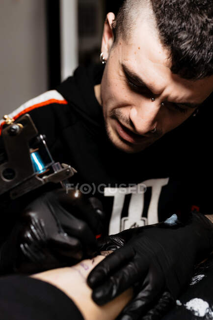 Stilvolle Mann mit Piercing mit Tätowiermaschine, um Tätowierung auf Bein der Ernte Kunden während der Arbeit im Salon zu machen — Stockfoto