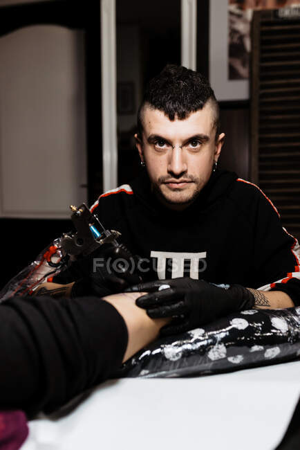 Stylish happy man with piercing looking at camera while using tattoo machine to make tattoo on leg of crop anonymous customer during work in salon — Stock Photo