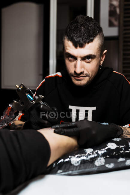 Homem feliz elegante com piercing olhando para a câmera ao usar a máquina de tatuagem para fazer tatuagem na perna do cliente anônimo durante o trabalho no salão — Fotografia de Stock