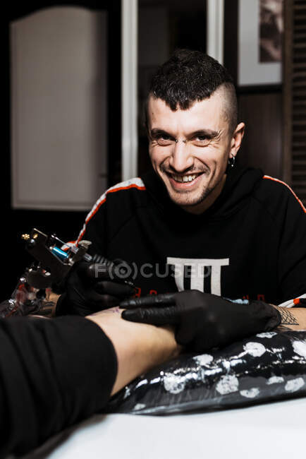 Elegante uomo felice con piercing guardando la fotocamera mentre usa la macchina del tatuaggio per fare tatuaggi sulla gamba del cliente anonimo del raccolto durante il lavoro nel salone — Foto stock