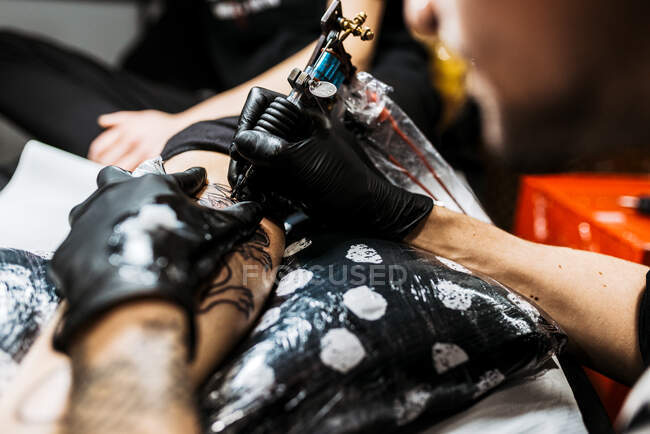 Dall'alto uomo irriconoscibile ritagliato con l'uso della macchina del tatuaggio per fare il tatuaggio sulla gamba del cliente del raccolto durante il lavoro nel salone — Foto stock
