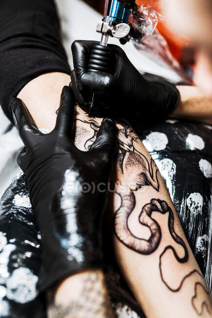De dessus recadré homme méconnaissable avec l'aide d'une machine à tatouer pour faire le tatouage sur la jambe du client de la culture pendant le travail dans le salon — Photo de stock