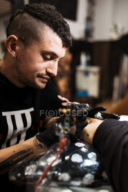 Стильний чоловік з пірсингом за допомогою татуювальної машини, щоб зробити татуювання на нозі клієнта врожаю під час роботи в салоні — стокове фото