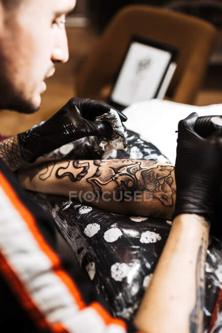 Сверху стильный мужчина с пирсингом с помощью тату-машины, чтобы сделать татуировку на ноге клиента урожая во время работы в салоне — стоковое фото