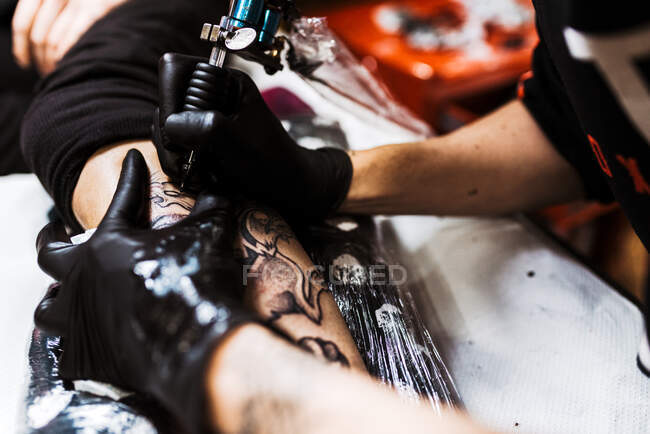 Свыше обрезанный неузнаваемый человек с помощью татуировки машины, чтобы сделать татуировку на ноге клиента урожая во время работы в салоне — стоковое фото