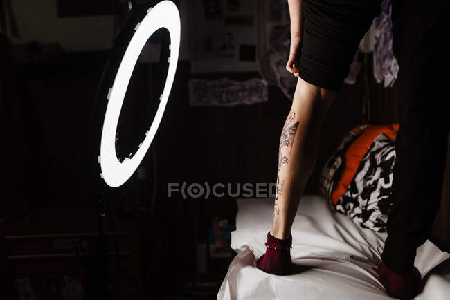 Cultivo irreconocible pierna de persona con tatuaje fresco en la lámpara brillante redonda durante la sesión de fotos en el salón de tatuajes - foto de stock