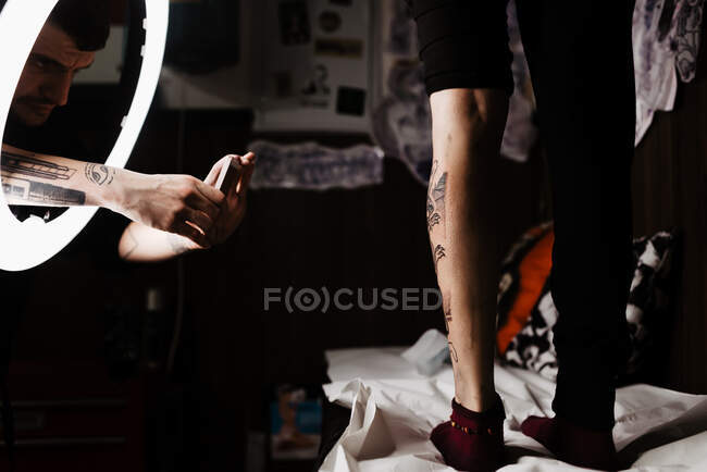 Hombre tatuador irreconocible usando teléfono inteligente para tomar la foto del tatuaje en la pierna del cliente de la cosecha para la cartera en el estudio contemporáneo - foto de stock