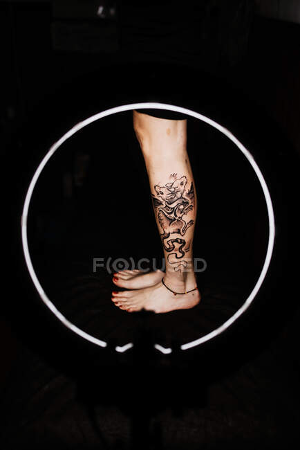 Cultivo pierna de mujer irreconocible con tatuaje fresco en lámpara brillante redonda durante la sesión de fotos en el salón de tatuajes - foto de stock