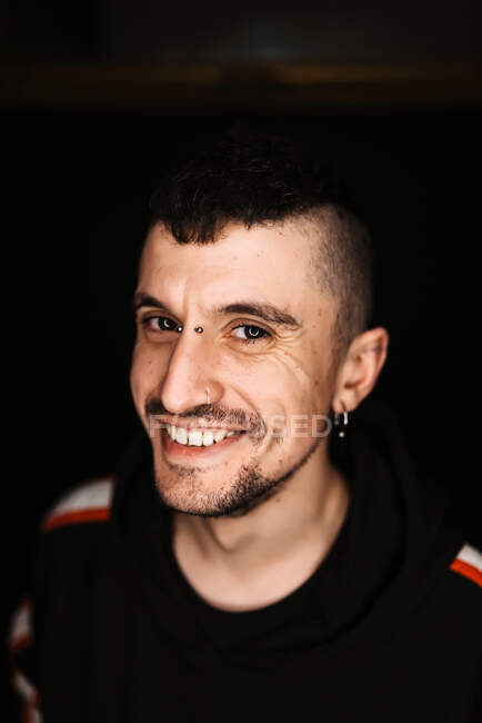Hombre adulto seguro con piercing y mohawk sonriendo y mirando a la cámara en la habitación oscura - foto de stock