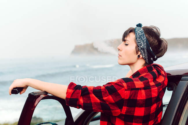 Side view of extraordinary woman with black short hair and bandana on head and piercing in red plaid shirt leaning on car and looking away at seaside — Stock Photo