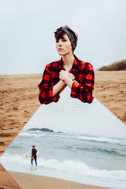Thoughtful sad young female in casual checkered shirt standing on sandy beach and holding large mirror with reflection of stormy sea and walking man — Stock Photo