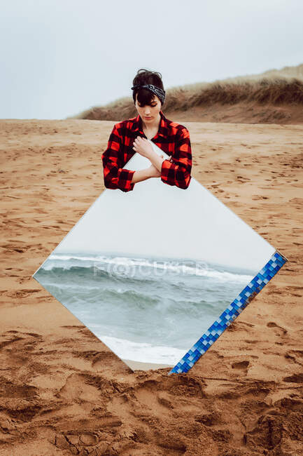 Щаслива самотня молода жінка в повсякденному вбранні стоїть на порожньому піщаному пляжі в похмурий день і тримає велике дзеркало з відображенням штормового моря — стокове фото