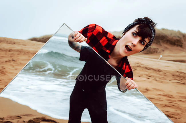 Удивленный шокирован молодой женщиной в повседневной одежде, стоящей на песчаном пляже и держащей большое зеркало с отражением урожая мужского пола против бурного моря — стоковое фото