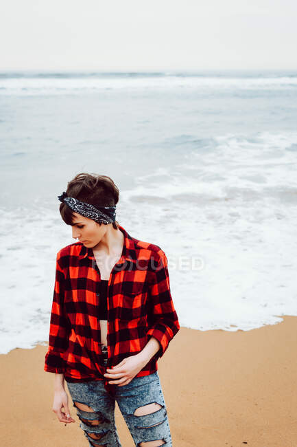 From above of pensive unhappy female in checkered shirt and ripped jeans standing with cigarette in hand on sandy beach with stormy sea in background — Stock Photo