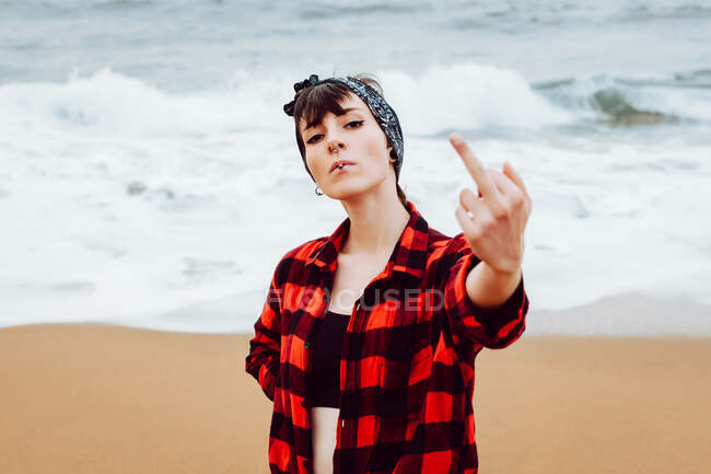 Высокомерная молодая женщина с пирсингом и сигаретой в расстегнутой рубашке, показывающая средний палец, стоя на песчаном пляже с морскими волнами на заднем плане — стоковое фото