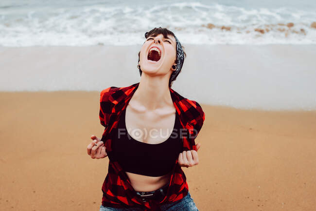 Mujer hipster joven irritado desesperado gritando en voz alta mientras se sienta en la playa de arena con olas de mar en el fondo - foto de stock