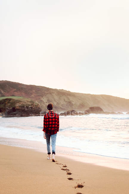 Rückansicht einer jungen kurzhaarigen Hipsterin in kariertem Hemd und Jeans, die am Strand spaziert und Fußspuren auf nassem Sand hinterlässt — Stockfoto