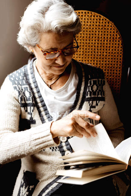Zufriedene Frau mit grauen Haaren und freundlichem Lächeln liest Buch am Fenster bei hellem Tag — Stockfoto