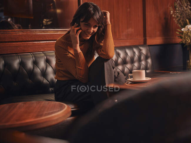 Stylish cheerful woman talking on mobile phone comfortably sitting at round table in cafe — Stock Photo