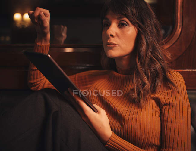 Elegante inteligente mulher surf tablet confortavelmente sentado com as pernas no sofá de couro preto no café — Fotografia de Stock