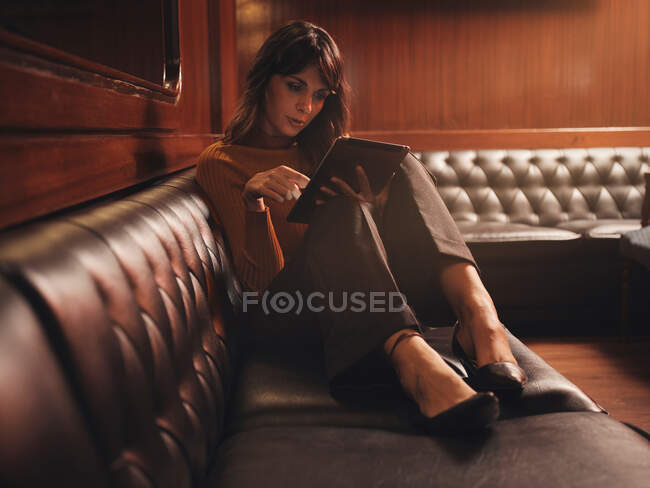 Стильна розумна жінка, серфінг планшет зручно сидить з ногами на чорному шкіряному дивані в кафе — стокове фото