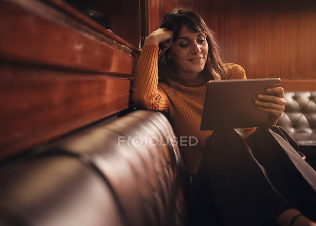 Élégant smart femme heureuse tablette de surf confortablement assis avec les jambes sur le canapé en cuir noir dans le café — Photo de stock