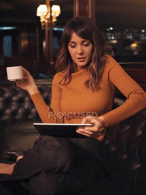 Élégant intelligent femme réfléchie surfer tablette confortablement assis sur le canapé en cuir noir dans le café en utilisant la tablette et boire du café dans une tasse — Photo de stock