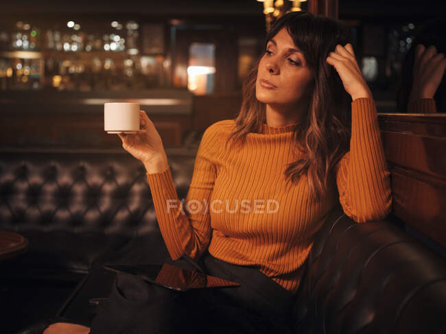 Elegante inteligente mulher pensativa surf tablet confortavelmente sentado no sofá de couro preto no café usando tablet e beber café em uma caneca — Fotografia de Stock