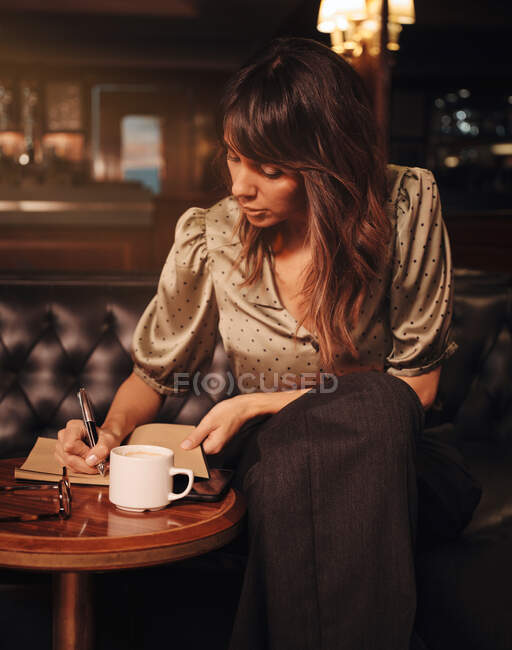 Mulher inteligente pensativa escrevendo com caneta em notebook em mesa redonda de madeira com caneca de café e óculos no café — Fotografia de Stock