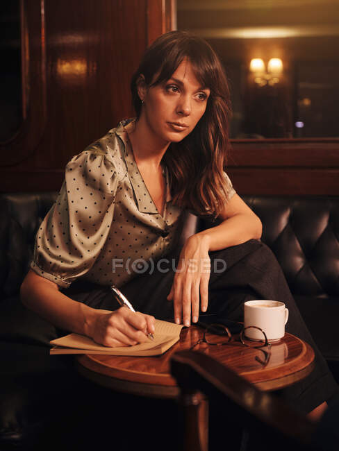 Thoughtful smart woman writing with pen in notebook at round wooden table with coffee mug and glasses in cafe — Stock Photo
