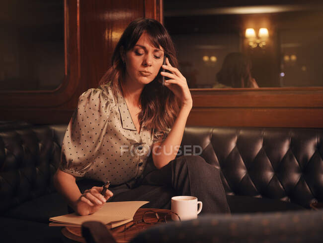 Стильна жінка уважно слухає інформацію на мобільному телефоні і пише в блокноті, комфортно сидячи за круглим столом в кафе — стокове фото