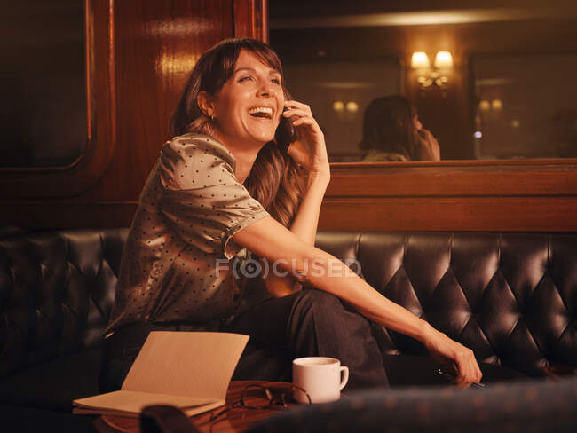 Elegante donna felice attentamente ascoltando le informazioni sul telefono cellulare e scrivendo nel notebook comodamente seduto a tavola rotonda nel caffè — Foto stock