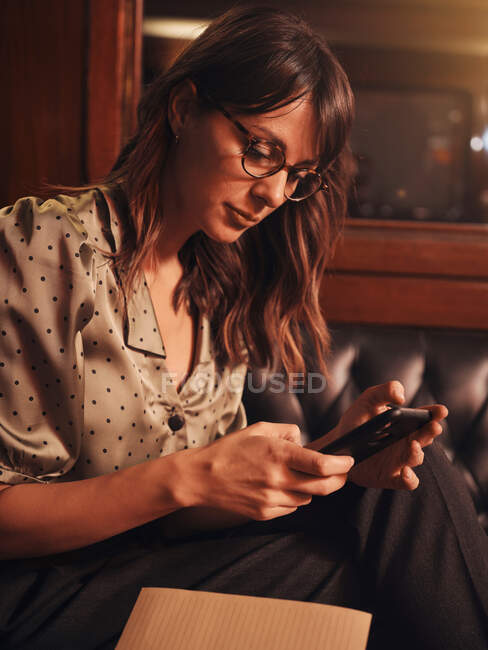 Side view of stylish thoughtful woman surfing mobile phone comfortable sitting at black leather sofa in cafe — Stock Photo