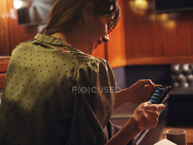 Вид збоку на стильну вдумливу жінку, що серфінгує мобільний телефон, зручно сидить на чорному шкіряному дивані в кафе — стокове фото