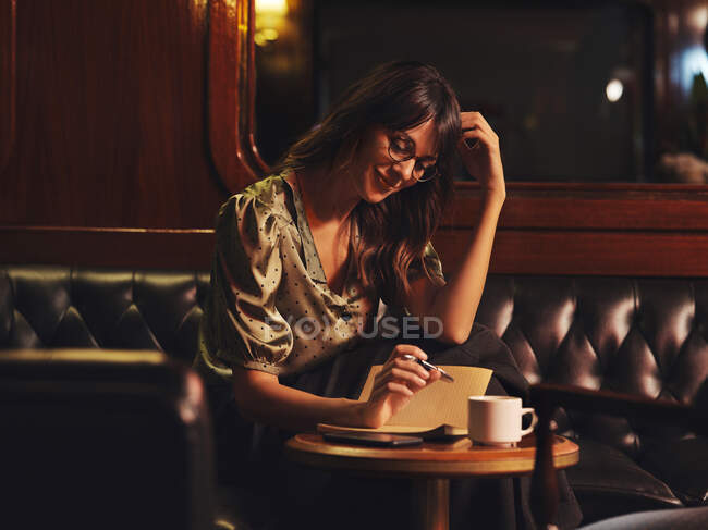 Pensive charming woman in glasses writing in notebook at round wooden table with coffee in cafe — Stock Photo