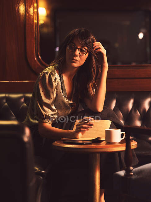 Donna affascinante pensieroso in bicchieri di scrittura in notebook a tavola rotonda in legno con caffè in caffè — Foto stock