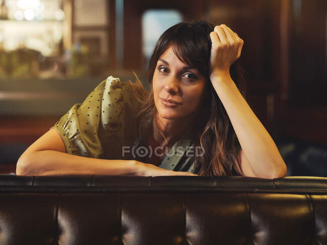 Вид сбоку усталой расслабленной женщины, опирающейся на спинку кожаного черного дивана и смотрящей на камеру в кафе — стоковое фото