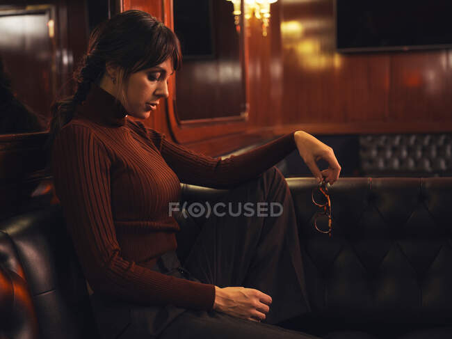 Femme élégante à la mode dans des vêtements à la mode assis sur un canapé en cuir noir et regardant loin — Photo de stock