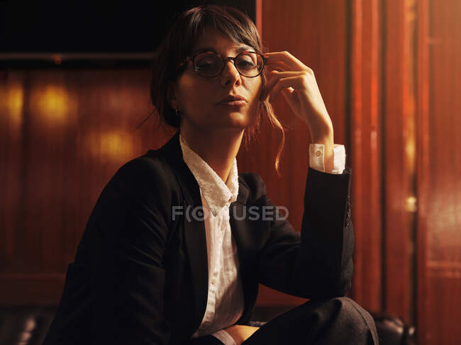 Femme élégante à la mode dans des vêtements et des lunettes à la mode assis sur un canapé en cuir noir et regardant la caméra — Photo de stock
