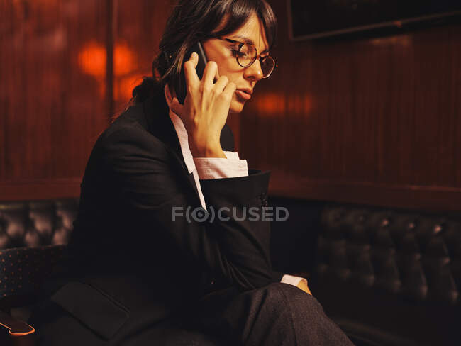 Elegante gerente de traje clásico hablando en el teléfono móvil cómodamente sentado en el sofá de cuero negro en la cafetería - foto de stock