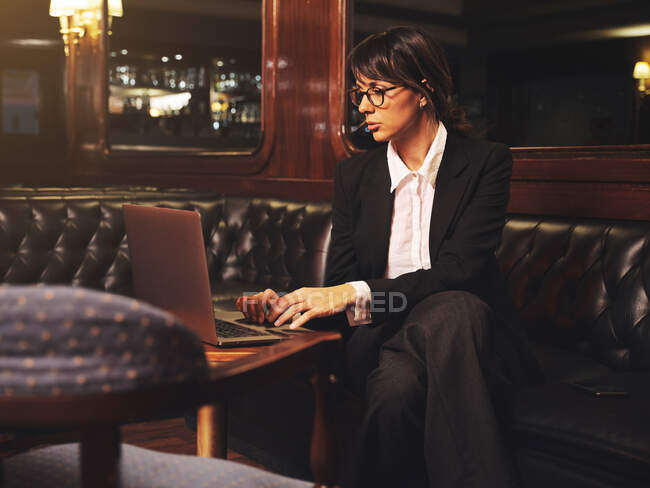 Smart business woman in glasses typing on laptop comfortably sitting in black leather sofa in cafe — Stock Photo