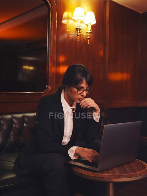 Mujer de negocios inteligente en gafas de escribir en el ordenador portátil cómodamente sentado en sofá de cuero negro en la cafetería - foto de stock
