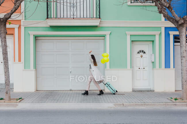 Side view of unrecognizable stylish female covering face with yellow balloons walking with suitcase on city street next to old styled colorful building — Stock Photo