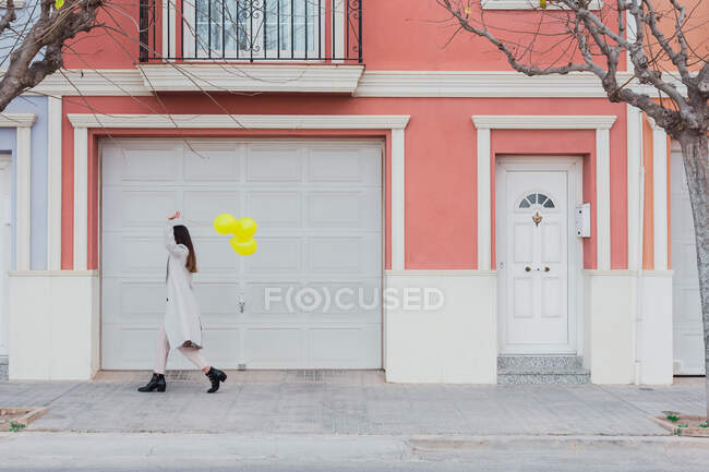 Vista laterale di irriconoscibile elegante volto femminile di copertura con palloncini gialli a piedi con valigia sulla strada della città accanto al vecchio edificio colorato in stile — Foto stock