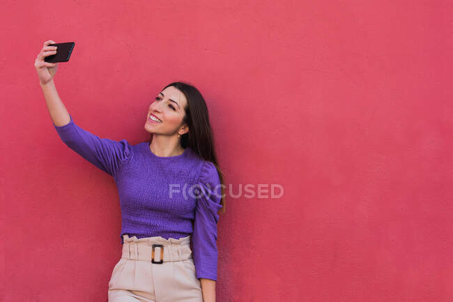 Jovem fêmea positiva em blusa violeta e calças bege-claras tirando uma selfie no celular enquanto estava de pé contra o colorido fundo da parede vermelha — Fotografia de Stock
