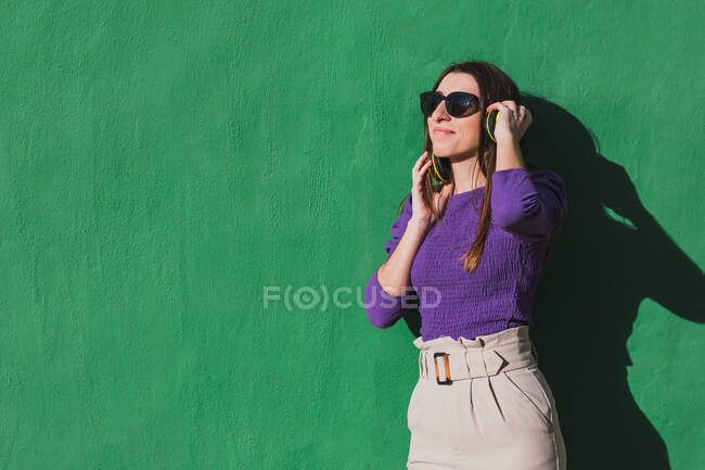 Positivo giovane femmina in camicetta viola e pantaloni beige chiaro parlando sul telefono cellulare mentre in piedi contro sfondo colorato parete verde — Foto stock