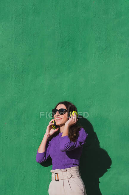 Jovem fêmea positiva em blusa violeta e calças bege claro falando no telefone celular, enquanto em pé contra fundo de parede verde colorido — Fotografia de Stock
