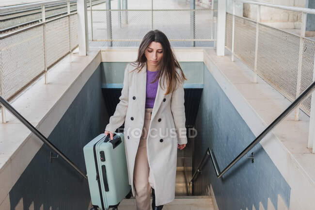 From above of elegant stylish young female traveler with suitcase walking up stairs at train station — Stock Photo