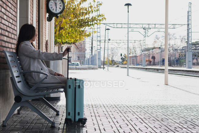 Vista lateral do jovem viajante elegante do sexo feminino com mala sentada no banco e esperando por transporte na estação ferroviária em dia ensolarado — Fotografia de Stock